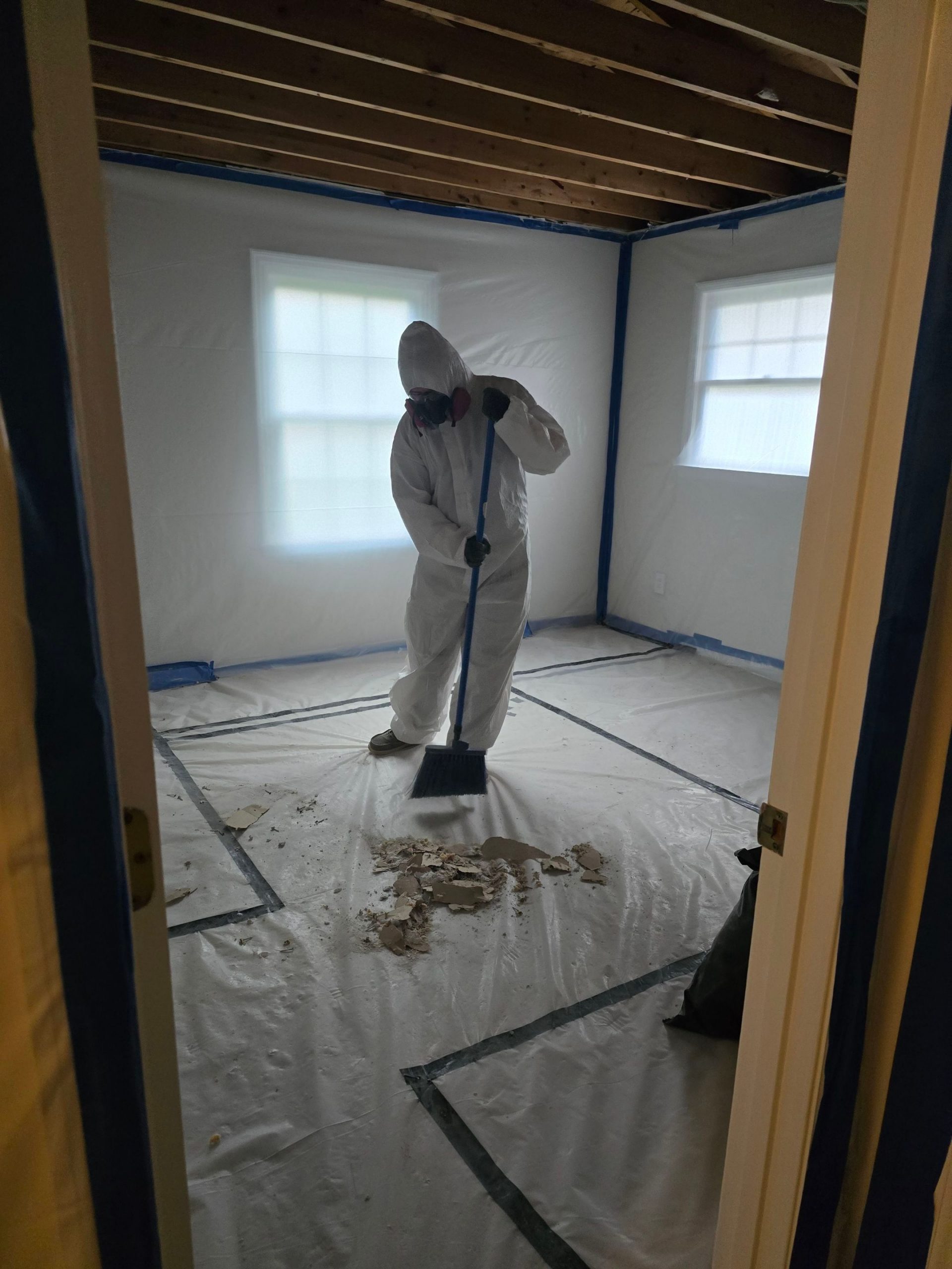 C2G Environmental technician in protective coveralls and respirator sweeping ceiling demolition debris inside a plastic-lined asbestos abatement containment area.