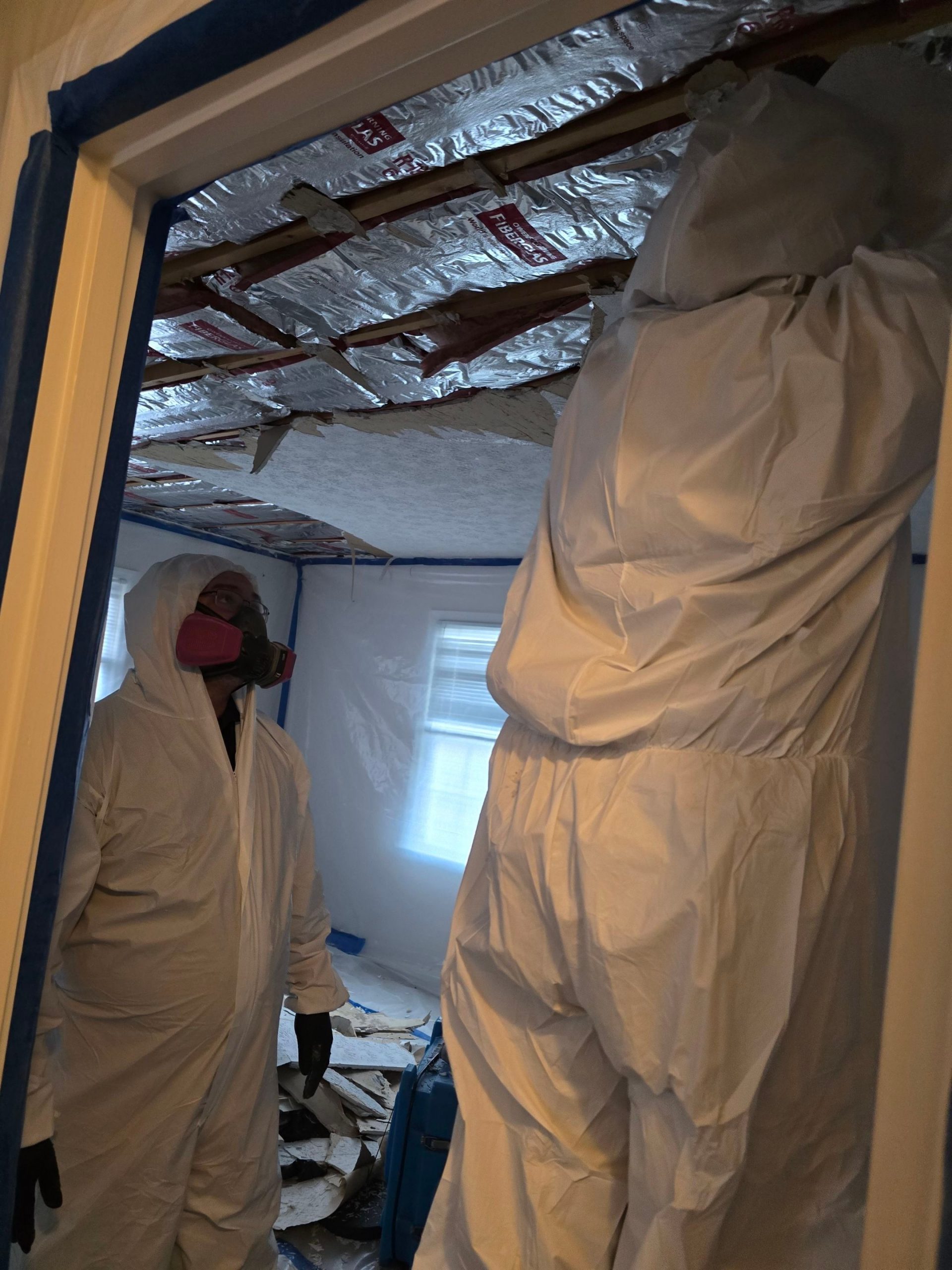 Two C2G Environmental technicians wearing protective suits and respirators removing ceiling materials inside a residential asbestos abatement containment area.
