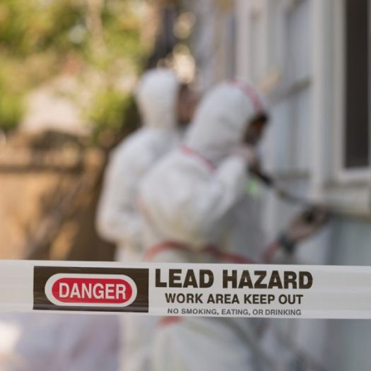 Lead hazard warning tape in front of workers wearing protective suits during exterior building work.
